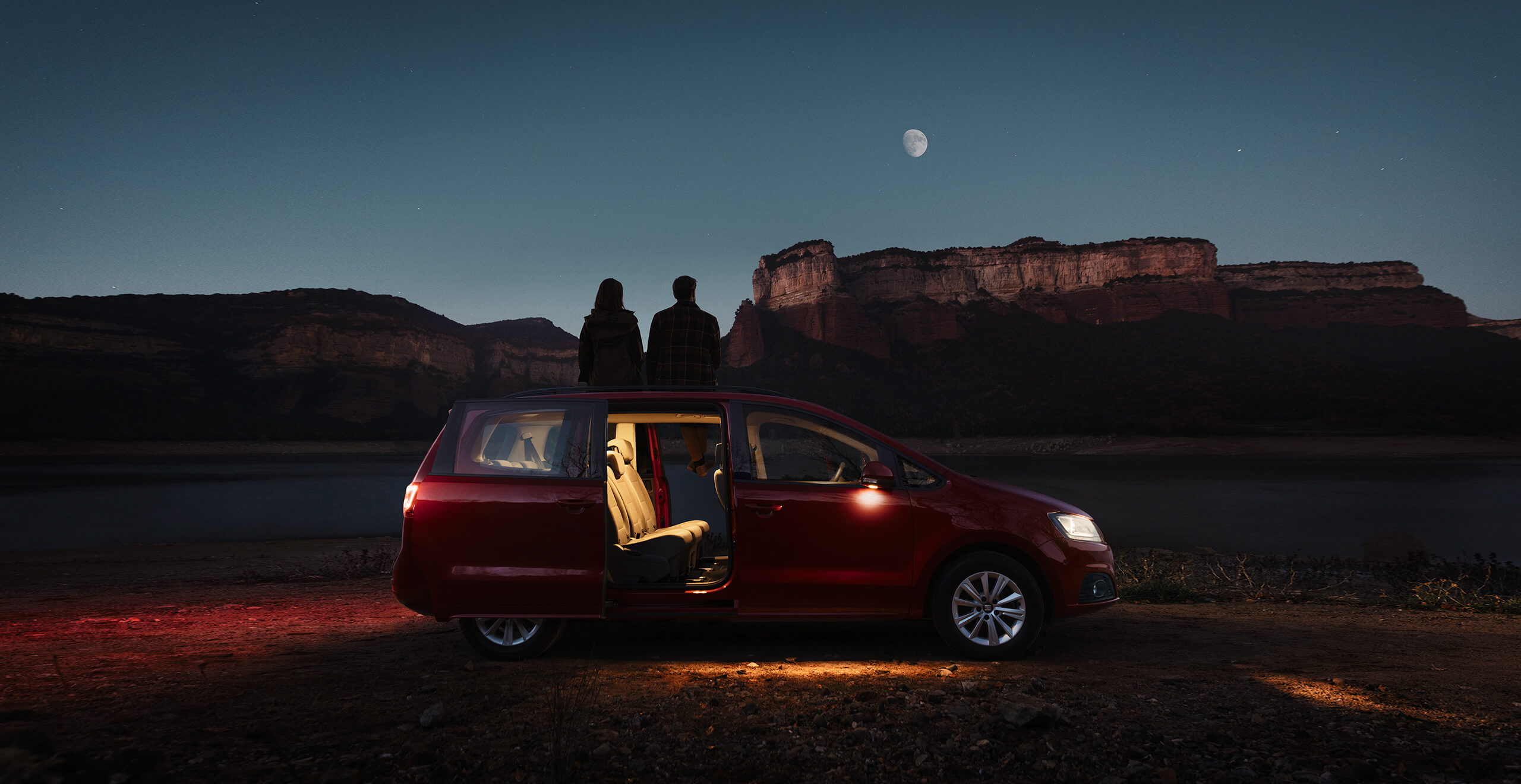 Dark night side view of SEAT Alhambra MPV with doors open and two people watching the moon and mountain landscape